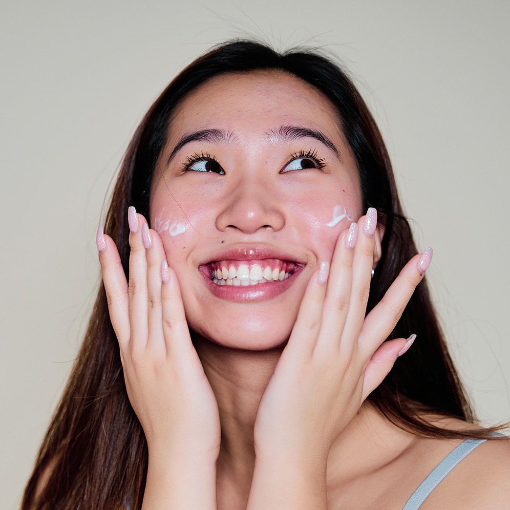 Front shot of woman smiling looking up as she applies moisturizer to both cheeks with both of her hands.